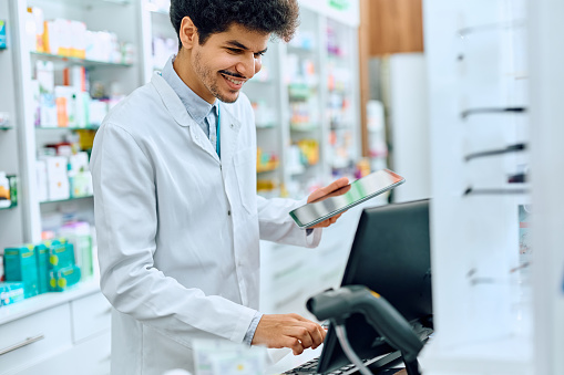 Happy Middle Eastern pharmacist using touchpad and desktop PC while working in a pharmacy.