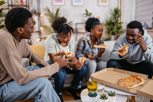 Group of friends sitting together and eating pizza in the living room at night.