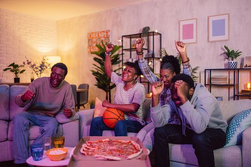 Black friends eating pizza and watching a basketball game in the living room.
