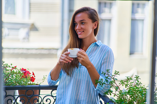 Portrait of a beautiful happy cute joyful smiling romantic woman with a aromatic coffee cup in hands in the morning on a balcony