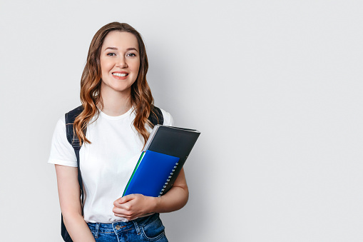 student girl wearing white t-shirt with backpack smiles and holds a notebook, copybook, folder in the hands isolated on dark white background