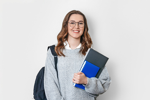 student girl dressed in a sweater with backpack and glasses smiles and holds a notebook, copybook, folder on a white background