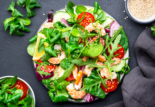 Tasty salad with avocado, shrimps, red tomatoes, cucumber, arugula, lamb lettuce, onion and sesame seeds on black table background. Top view, copy space