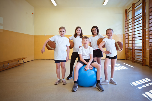 Elementary school girls and boy standing in gym, holding basketball balls and smiling at camera.