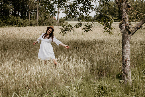 Woman in white dress and with hat on dancing in the field. There is a tree behind her.