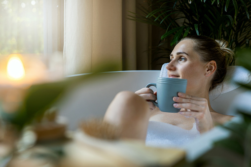 Young woman relaxing in the beautiful vintage bath full of foam in the retro bathroom decorated with candles