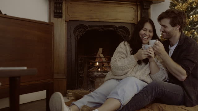 Coffee, relax and couple by fireplace on Christmas laughing at funny comedy. Tea, xmas and happy man and woman on holiday drinking beverage, love and enjoying quality time together on floor in home