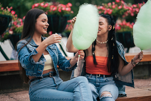 Two female friends eating cotton candy and siting at the bench. Looking happy.