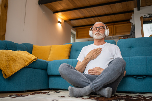 Mature senior man sitting on floor practicing guided meditation at home, relaxing body and mind concept