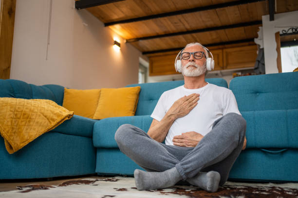 Mature senior man sitting on floor practicing guided meditation at home, relaxing body and mind concept stock photo