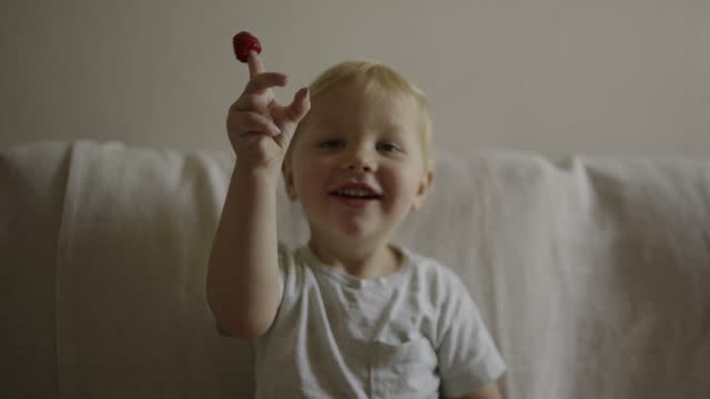Happy, cute and baby boy eating a raspberry for a healthy, organic and nutrition snack at home. Smile, sweet and toddler child enjoying red fruit for health and wellness on a sofa in the living room.