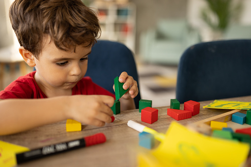 Cute little boy playing with toy blocks while sitting at the table at home.