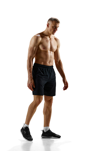 Full-length portrait of handsome young man with relief, fit, strong, ,muscular body standing shirtless against white studio background. Concept of sport, competition, health, strength and speed, ad