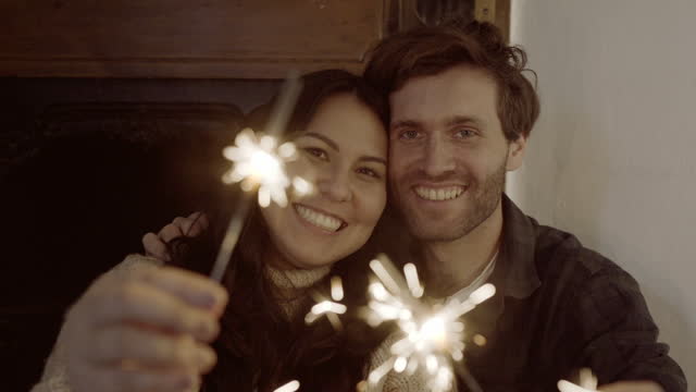 Young couple, sparkle firework and Christmas celebration for smile on face, excited or culture in home living room. Man, woman and happy for xmas tradition, light or play with love on festive holiday