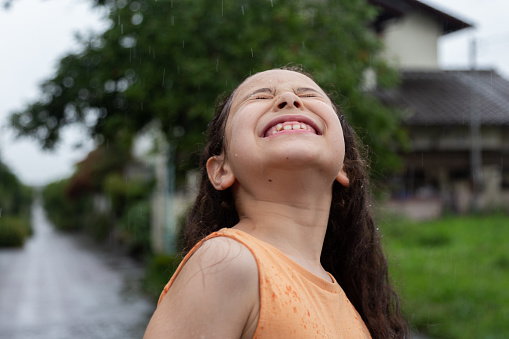 Young girl with smile is enjoying herself in the rain. Close-up of girl under the summer rain. Portrait of girl outdoors.