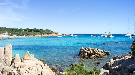 In the photograph, a stunning beach in Sardinia can be seen. The crystal-clear turquoise waters are gently lapping at the white sandy shore, with a few scattered rocks and greenery adding to the natural beauty of the setting. The bright sun is shining down upon the beach, casting a warm glow and creating sharp shadows on the sand. In the distance, the cerulean sea meets the cloudless sky, making for a breathtaking panoramic view. The entire scene is peaceful, serene, and inviting, making it the perfect destination for anyone seeking a relaxing and picturesque vacation.