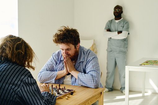 Young nervous man keeping palms put together by face and looking at chessboard while playing with other male patient of mental hospital
