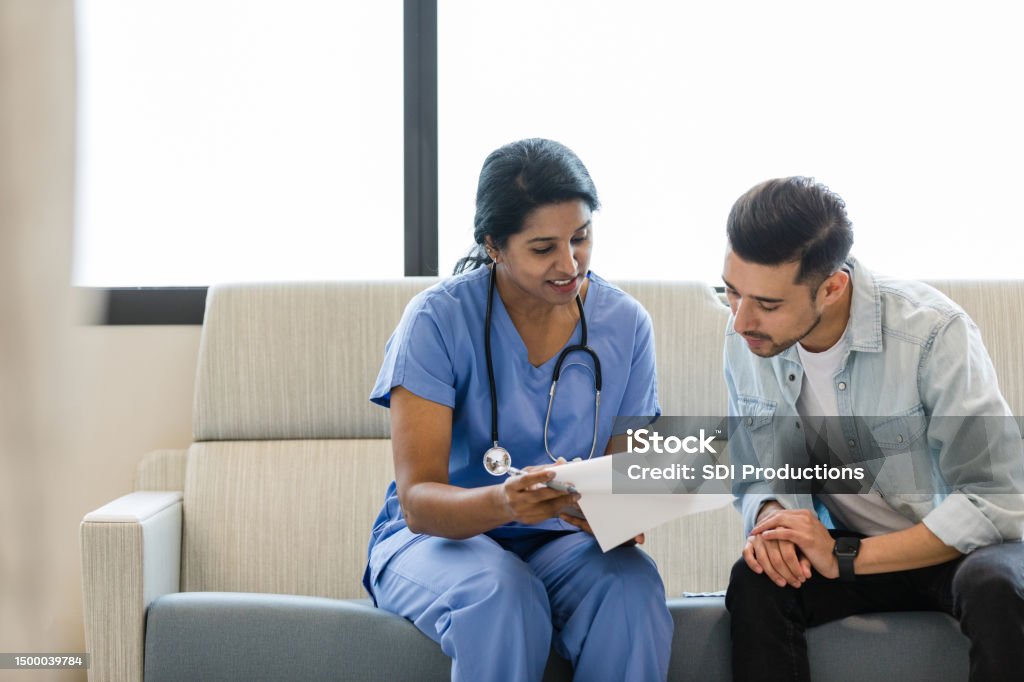 Young man and female doctor review the medical chart together The young man and female doctor sit on the hospital room couch and review the medical chart together. Doctor Stock Photo