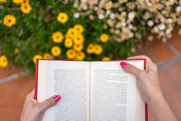 Woman's hands on a book while reading in the garden next to flowering plants