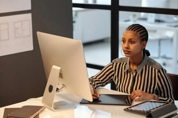 Young African American webdesigner or artist with stylus drawing sketch on touchpad screen while sitting by workplace in front of computer