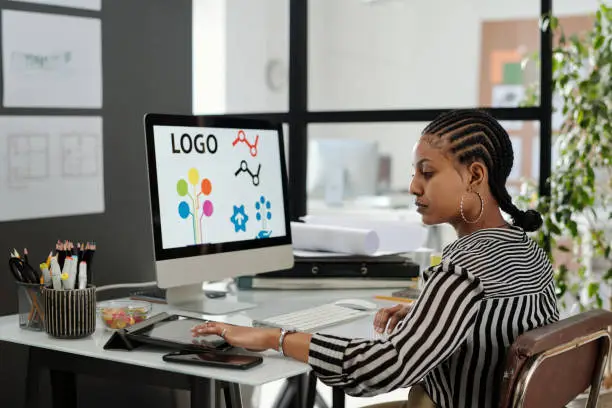 Side view of young serious African American female webdesigner sitting by workplace in front of computer monitor and using tablet