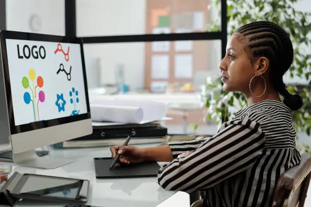 Serious African American female webdesigner sitting by desk in front of computer monitor with logo on screen and working over new project