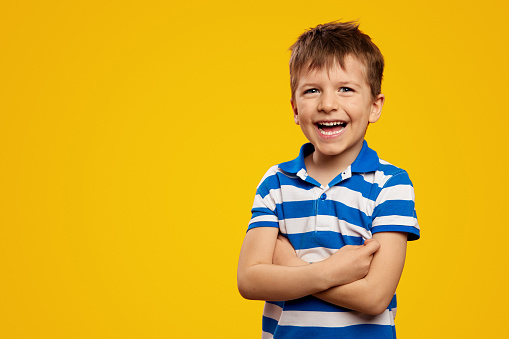 Portrait photo of adorable kid boy wearing blue striped polo, laughing while standing with arms crossed against yellow background with empty space