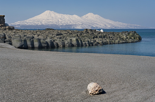 Russia. Far East, Kuril Islands. View of the Okhotsk coast of Iturup island with ash sand of volcanic origin against the background of two snow-covered volcanoes Chirip and Bogdan Khmelnitsky.