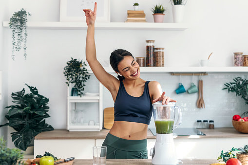 Shot of athletic woman preparing smothie while singing, dancing and listening music with earphones in the kitchen at home.