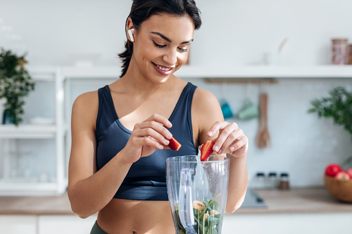 Shot of athletic woman preparing smothie with vegetables and fruits while listening music with earphones in the kitchen at home.