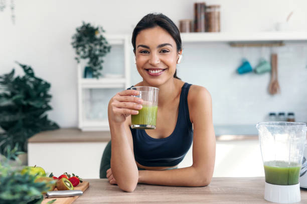 donna sorridente atletica che beve smothie mentre ascolta musica con gli auricolari in cucina a casa. - usare la bocca foto e immagini stock