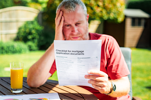 Color image depicting a mid adult man reading a checklist for mortgage application documents. The man is sitting at a chair and table outside in his garden and he has a worried expression on his face.