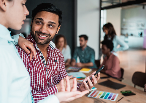 Portrait of a handsome ethnic businessman standing in front of his team in board room. Young businessman holding smart phone and talking with female coworker. Multi-ethnic group of creative people on a business meeting in modern office.