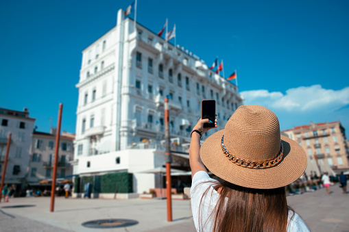 Young woman tourist with sun hat take a photo of Cannes, France. Using phone to photographing European city. Copy space