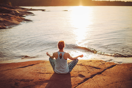 Young adult woman meditating at lake shore at sunset