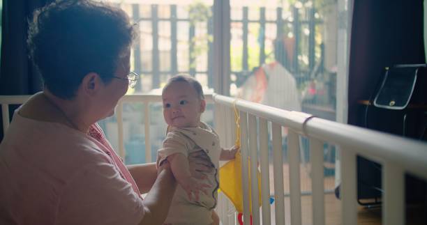 felice nonna anziana donna asiatica che insegna tenendo in braccio la sua nipote bambino imparare a stare in piedi fa i primi passi a casa, due generazioni di età vecchio nonno di famiglia che si gode l'attività insieme - kii foto e immagini stock