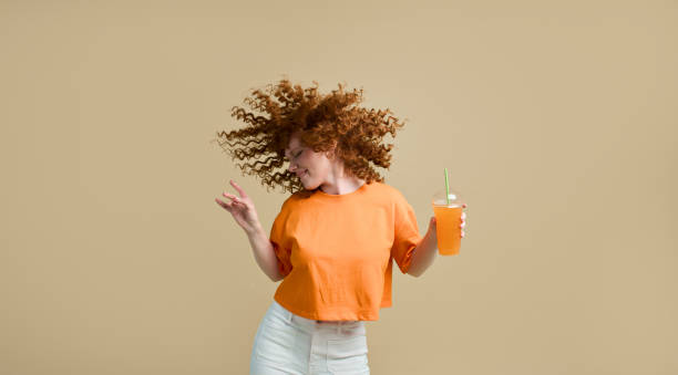 Happy carefree young wavy-haired ginger woman dancing holding fresh juice drink cocktail in hand having fun celebrating start of relax vacation time isolated on turquoise copy space background stock photo