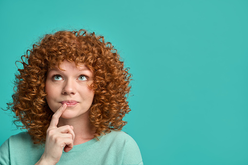 Young redhead woman hipster teenager with curly hair wearing casual clothes over turquoise background thinking deeply about something, keeping index finger near lips, looking upward copy space