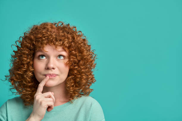 Headshot portrait of thoughtful pensive young ginger woman with curly hair holding finger on lips looking upward against turquoise studio wall background with copy space for text advertisement stock photo