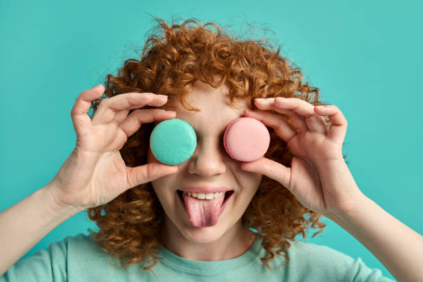 Headshot portrait of funny cute young ginger girl with curly hair holding tasty macaroon sweet cakes in front of eyes showing tongue making joke and fun isolated on green studio wall background stock photo