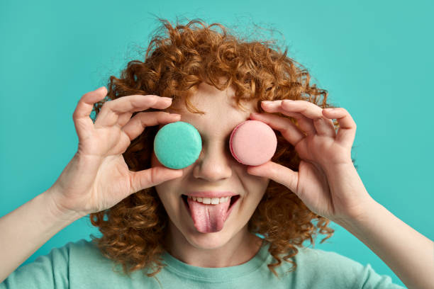 Headshot portrait of funny cute young ginger girl with curly hair holding tasty macaroon sweet cakes in front of eyes showing tongue making joke and fun isolated on green studio wall background Headshot portrait of cheerful positive little teenager ginger girl covering eyes with dessert macaroon cakes isolated on green color studio wall background. Beautiful woman enjoying sweet biscuits eye candy stock pictures, royalty-free photos & images
