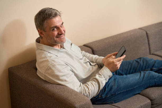 istock Smiling male holding phone and sitting on couch 1500070163