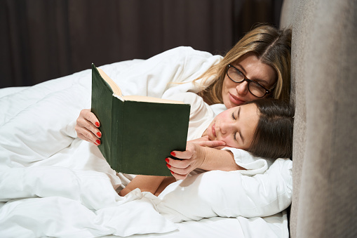 Mother in glasses lying near sleeping girl and looking into book in the bedroom
