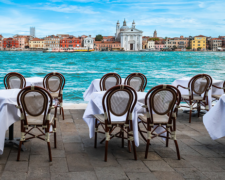 Tables with tablecloth and wooden chairs along the Giudecca canal in Venice. Religious church and colorful buildings in the background.