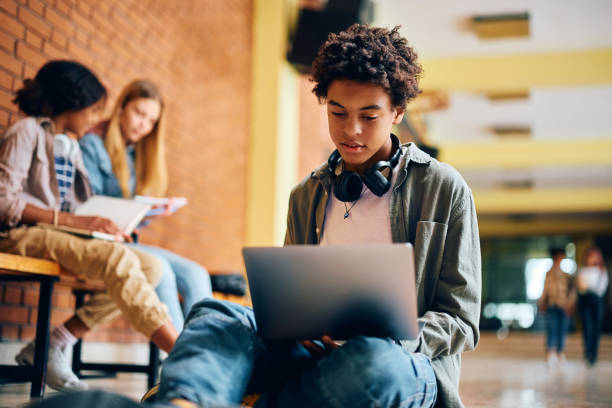 Black high school student surfing the net on laptop in a hallway.
