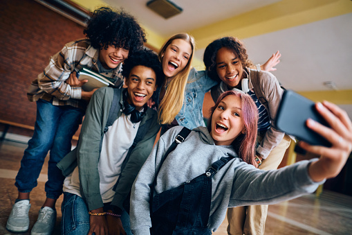 Group of happy high school students having fun while taking selfie with cell phone in hallway.