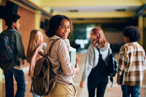 Happy black teenage girl in high school hallway looking at camera. Happy African American high school student walking through hallway with her friends and looking at camera. school building stock pictures, royalty-free photos & images