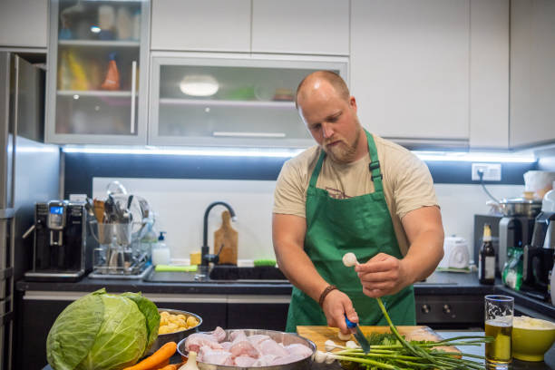 a blond man with a beard and a green apron prepares dinner in his home kitchen. - routine foods and drinks clothing household equipment imagens e fotografias de stock