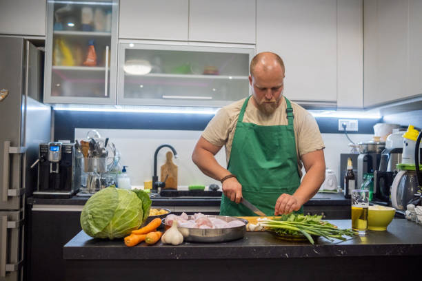 a blond man with a beard and a green apron prepares dinner in his home kitchen. - routine foods and drinks clothing household equipment imagens e fotografias de stock