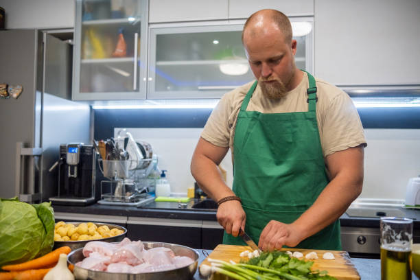 a blond man with a beard and a green apron prepares dinner in his home kitchen. - routine foods and drinks clothing household equipment imagens e fotografias de stock